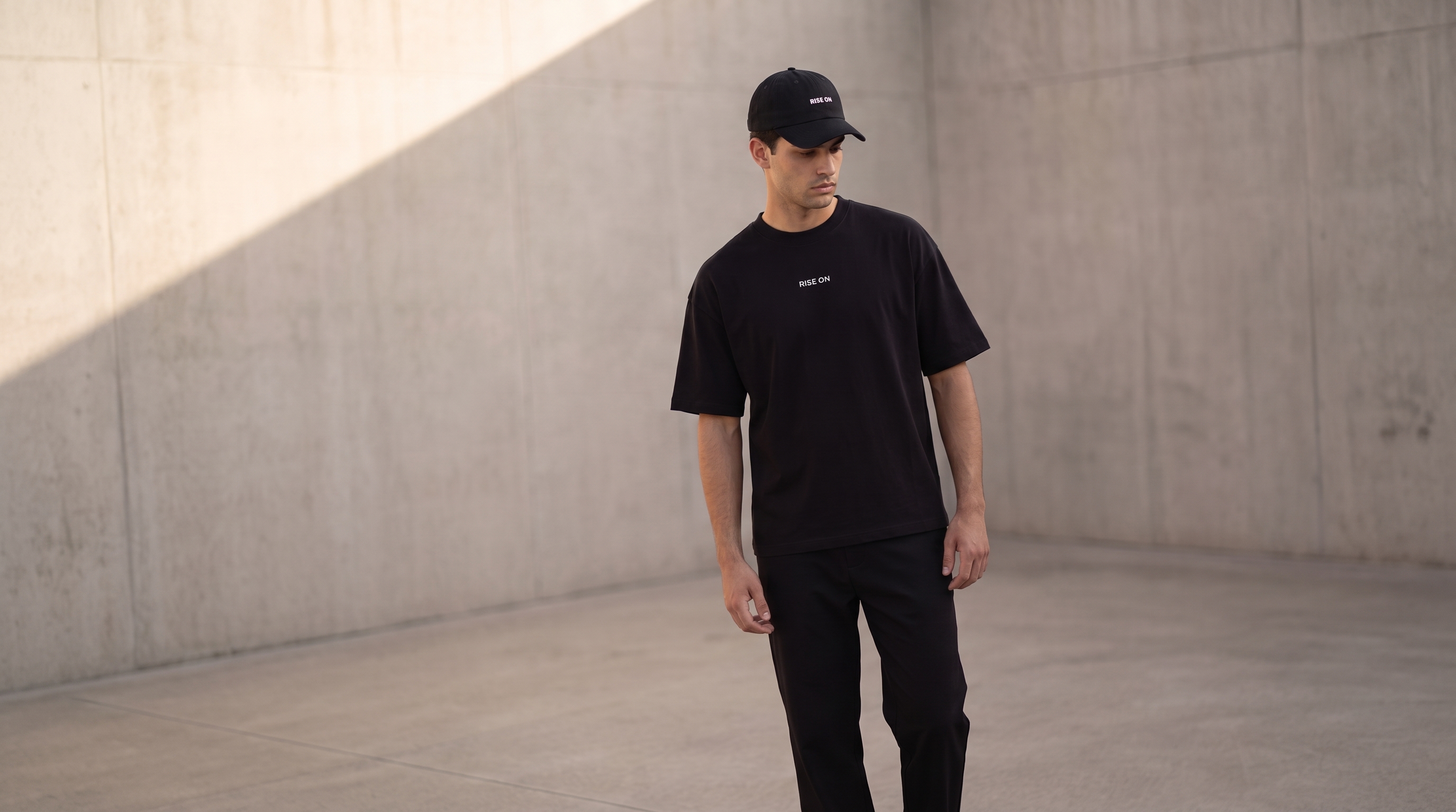 Modelo RISE ON con camiseta negra y gorra en un entorno editorial de hormigón